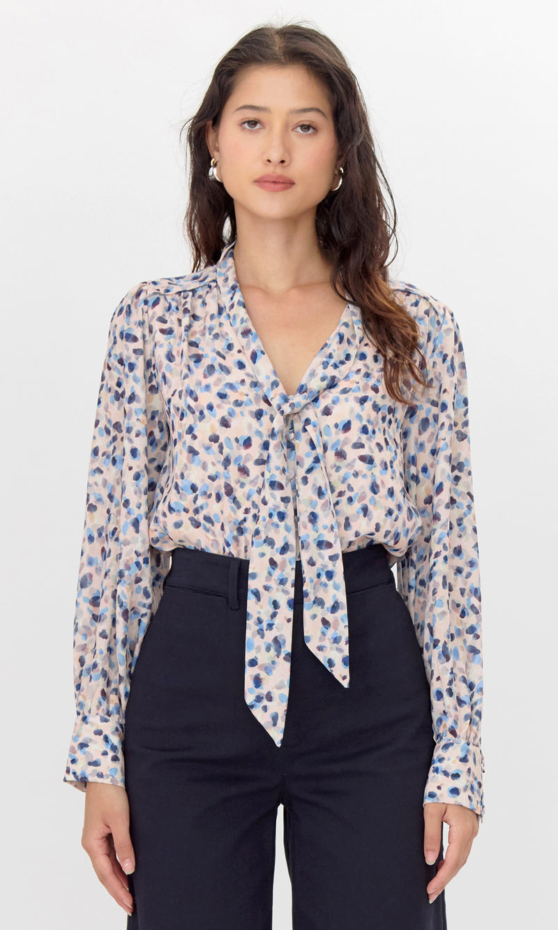 Female model wearing pink, tie-neck blouse with blue polka-dots. The blouse is tucked into her trousers.