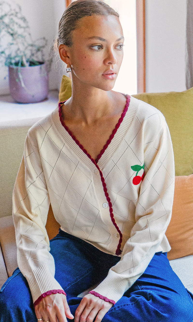 Model sits on a couch wearing a knit cardigan with a cherry design on the chest. The cardigan shows a white button closure and a red trimming along the neckline and sleeves.